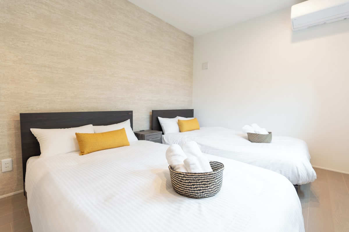 A hotel room with two beds, each with white bedding and a single yellow pillow. There is a woven basket on the bed in the foreground containing folded towels.