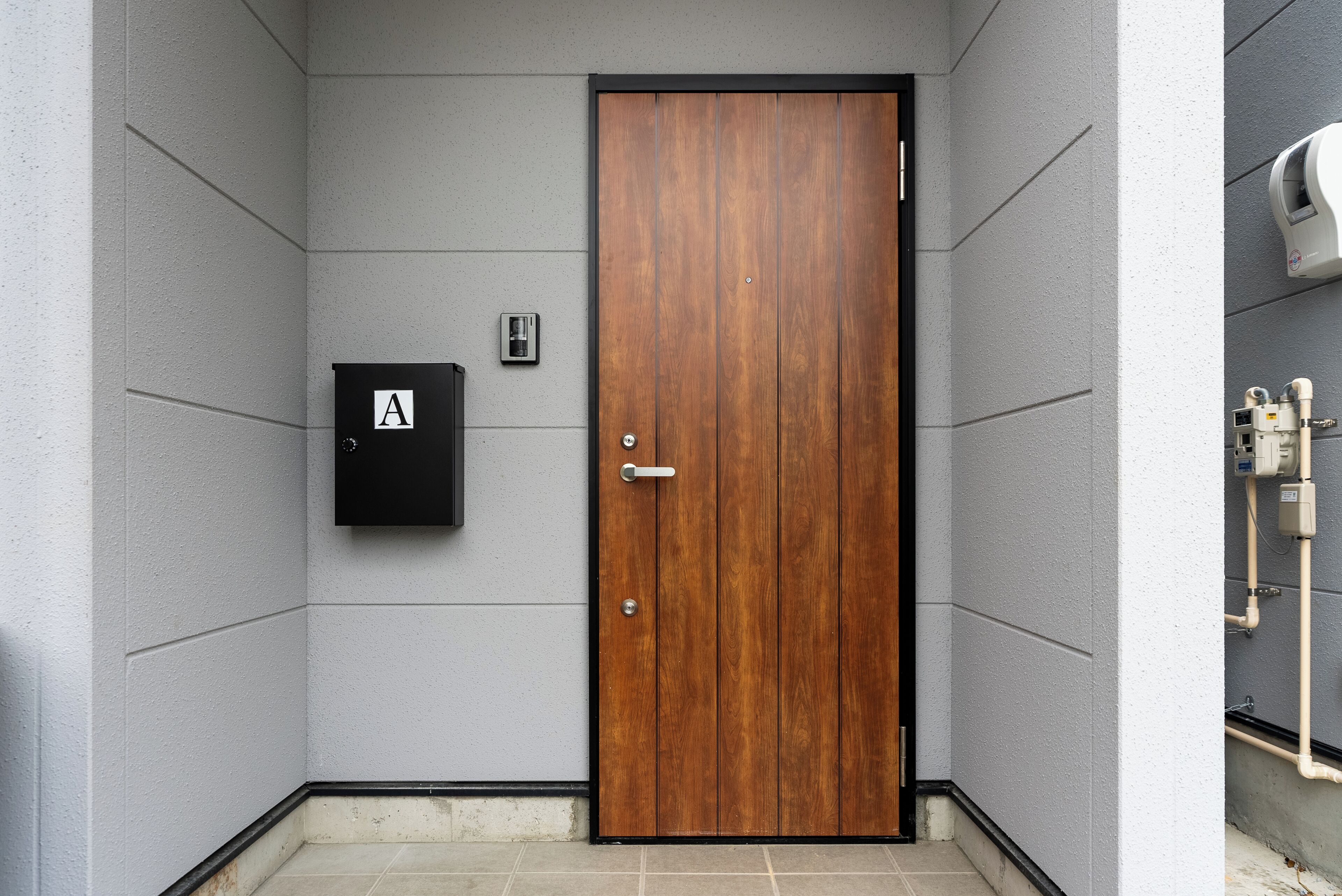 Una puerta de madera con un picaporte negro, un buzón negro con la letra 'A' y una pequeña caja eléctrica en la pared.