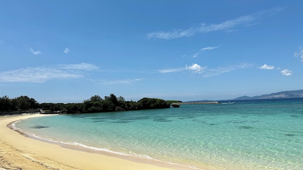 A beach with clear turquoise water, a sandy shore, and a distant view of land with vegetation and a boat. - Grandioso Okinawa Pool Villa Onna 13 (Onna)