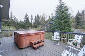 Une terrasse en bois avec un jacuzzi, des chaises blanches et une vue sur les arbres.