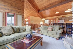 A cozy cabin interior with a wooden ceiling, two sofas, a coffee table, and a dining area with chairs and a table.