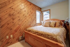 A bedroom with a wooden sloped ceiling, a bed with patterned bedding, and a window with a view of a building. - Alaskan Cabin Escape w a Hot Tub (Anchorage)