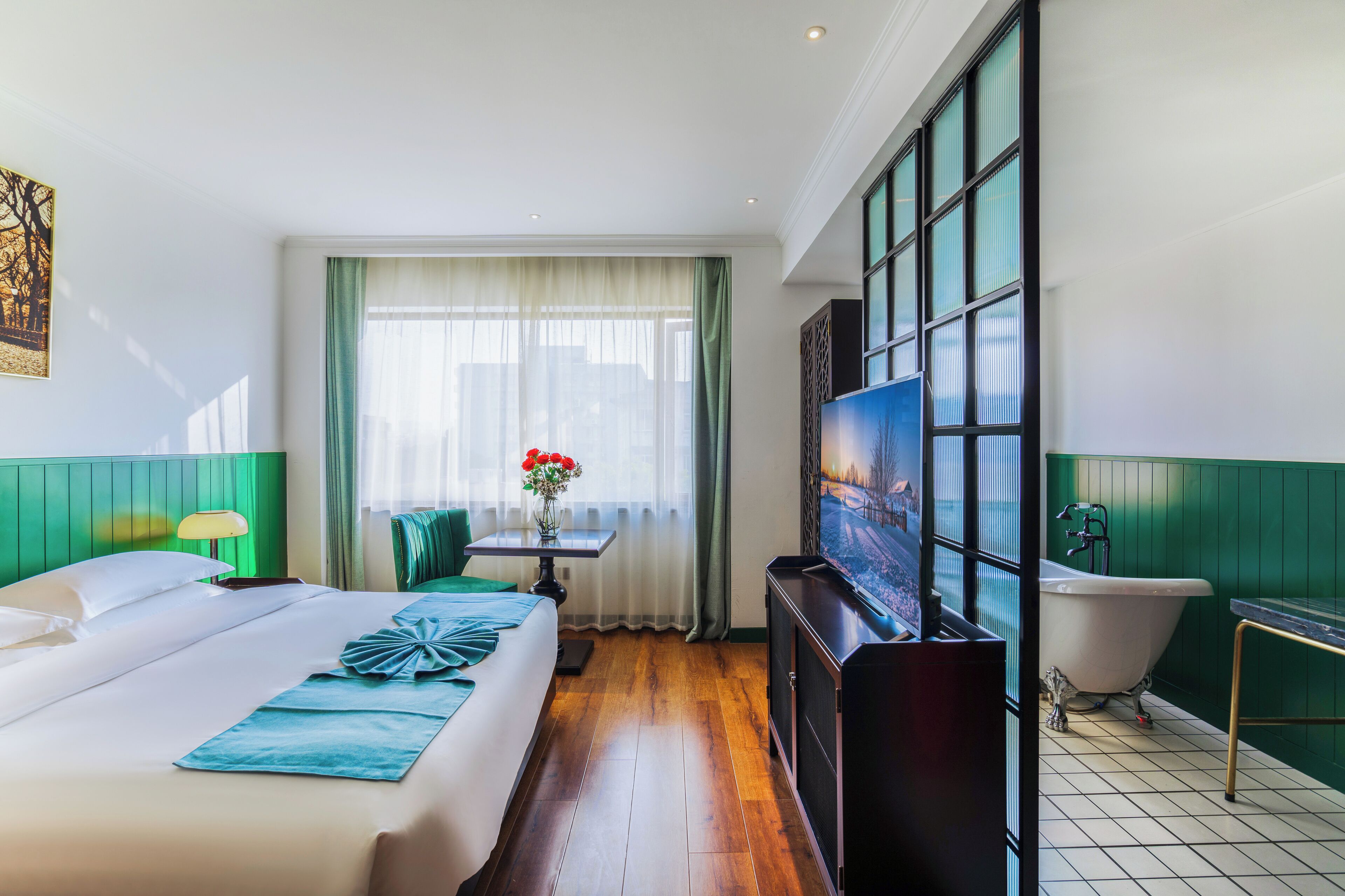 A hotel room with a bed, a table with a vase of flowers, a large window with curtains, a bathtub, and a television.