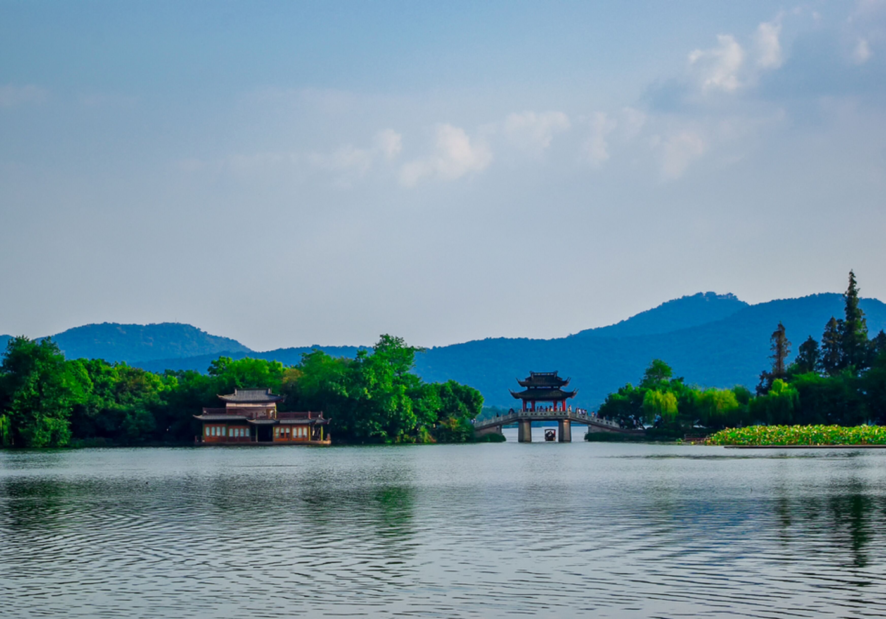 A traditional Chinese pavilion over a lake, surrounded by lush greenery and mountains in the background.