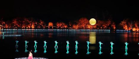 A nighttime scene with a large illuminated moon, a reflective water body, and a traditional building with red foliage in the background.