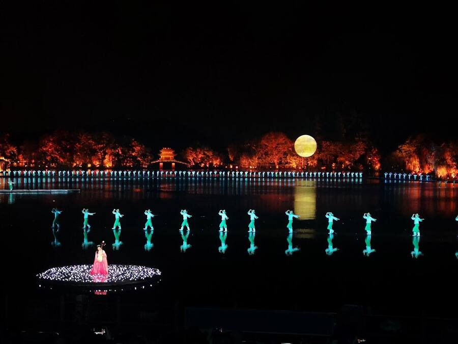 A nighttime scene with a large illuminated moon, a reflective water body, and a traditional building with red foliage in the background.