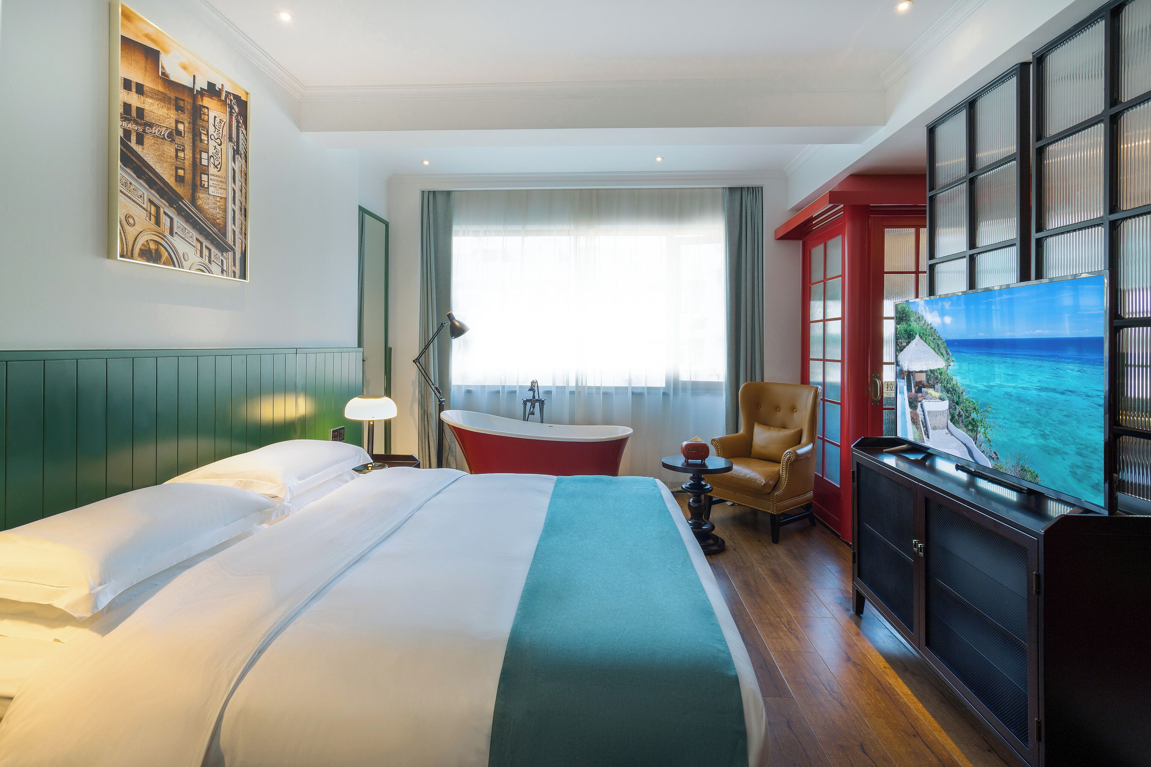 A hotel room with a large bed, a red bathtub, a chair, a TV, and a painting on the wall.