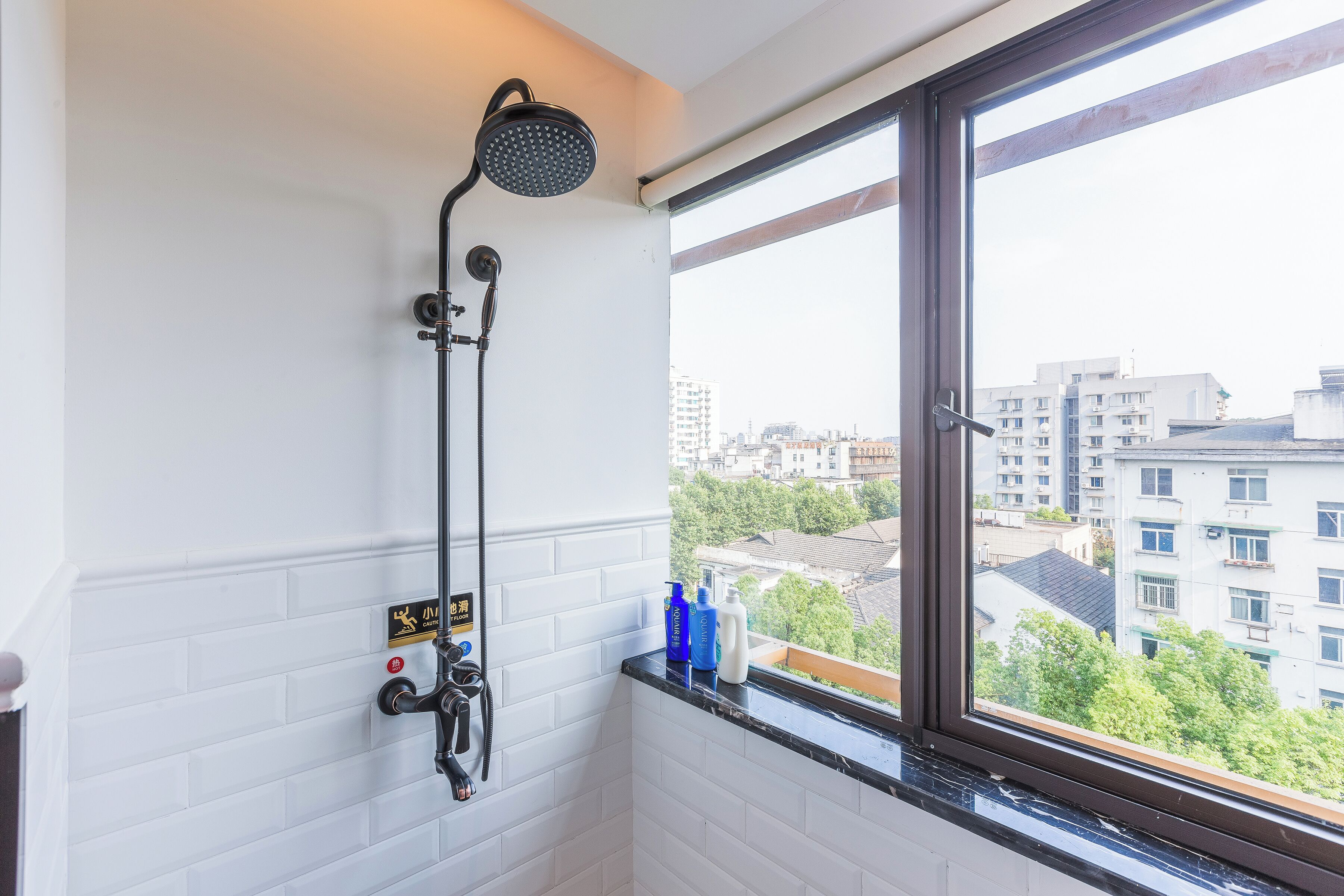A bathroom with a shower, a window with a city view, and toiletries on the windowsill.
