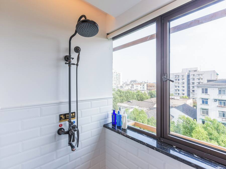 A bathroom with a shower, a window with a city view, and toiletries on the windowsill.