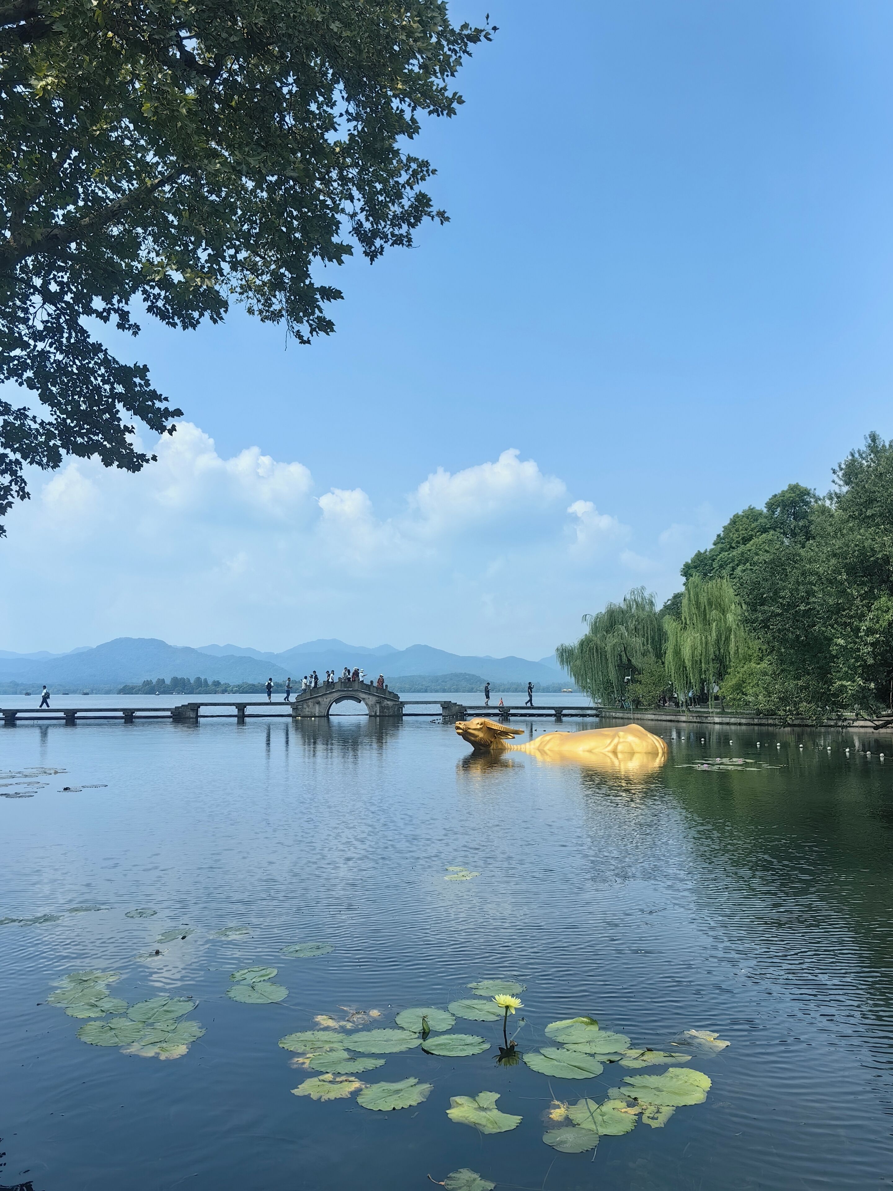 A serene lake with a stone bridge, lotus flowers, and a statue of a golden creature.