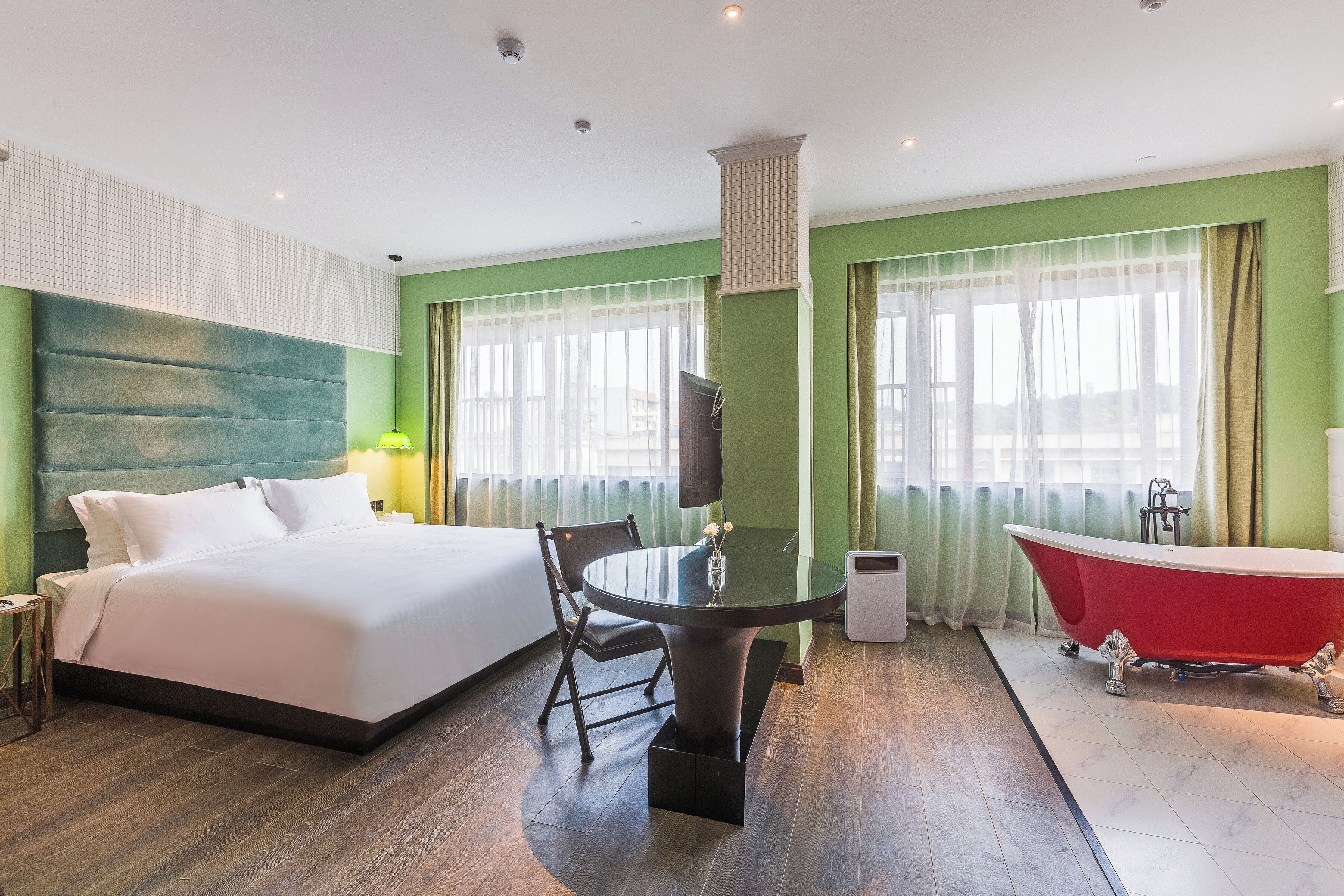 A hotel room with a bed, a round table with chairs, a red bathtub, and large windows with curtains.