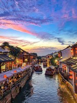 Um canal estreito ladeado por edifícios tradicionais, lanternas vermelhas, barcos de madeira e áreas de estar ao ar livre.