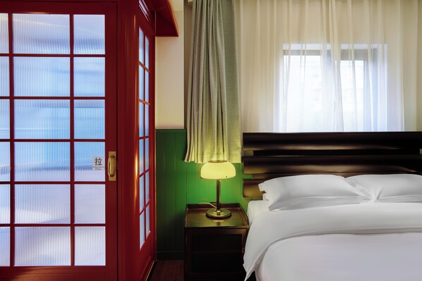 A hotel room with a bed, a nightstand with a lamp, a window with curtains, and a door with a pull handle.