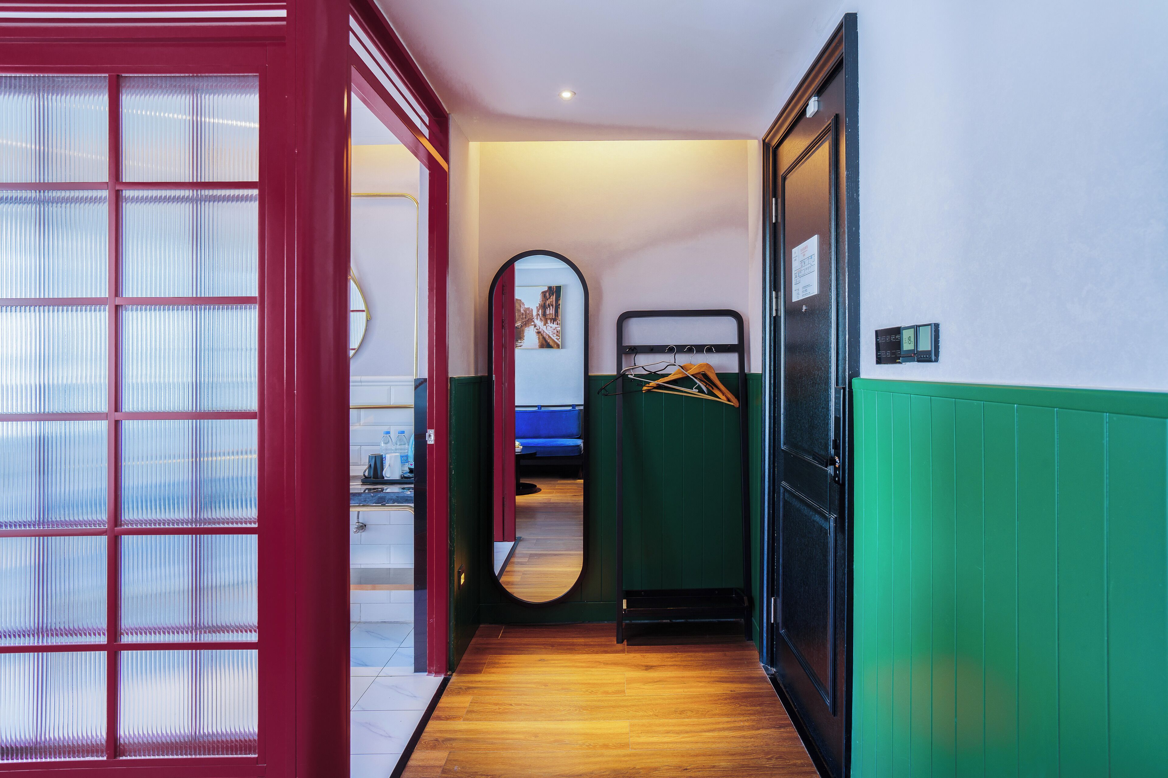 Un pasillo estrecho con suelo de madera, una puerta de vidrio con marco rojo, una pared verde y una cama con cabecera azul.