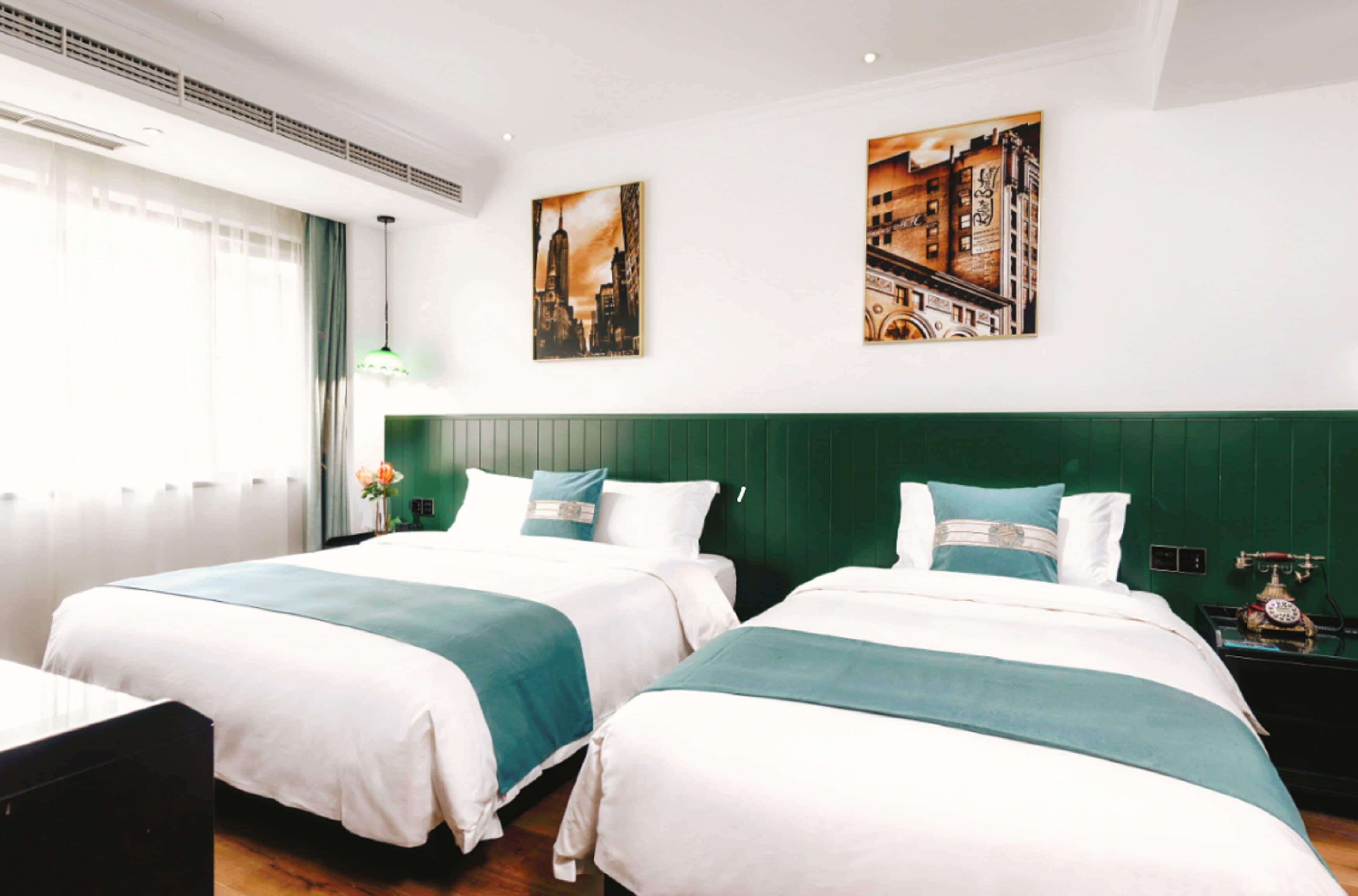 A hotel room with two beds, a green headboard, a window with curtains, and two paintings on the wall.