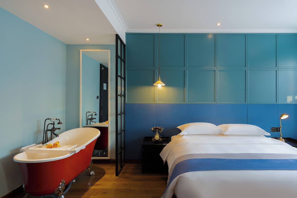 Una habitación de hotel moderna con una bañera de patas de león roja, una cama grande con ropa de cama blanca y una pared de acento azul.