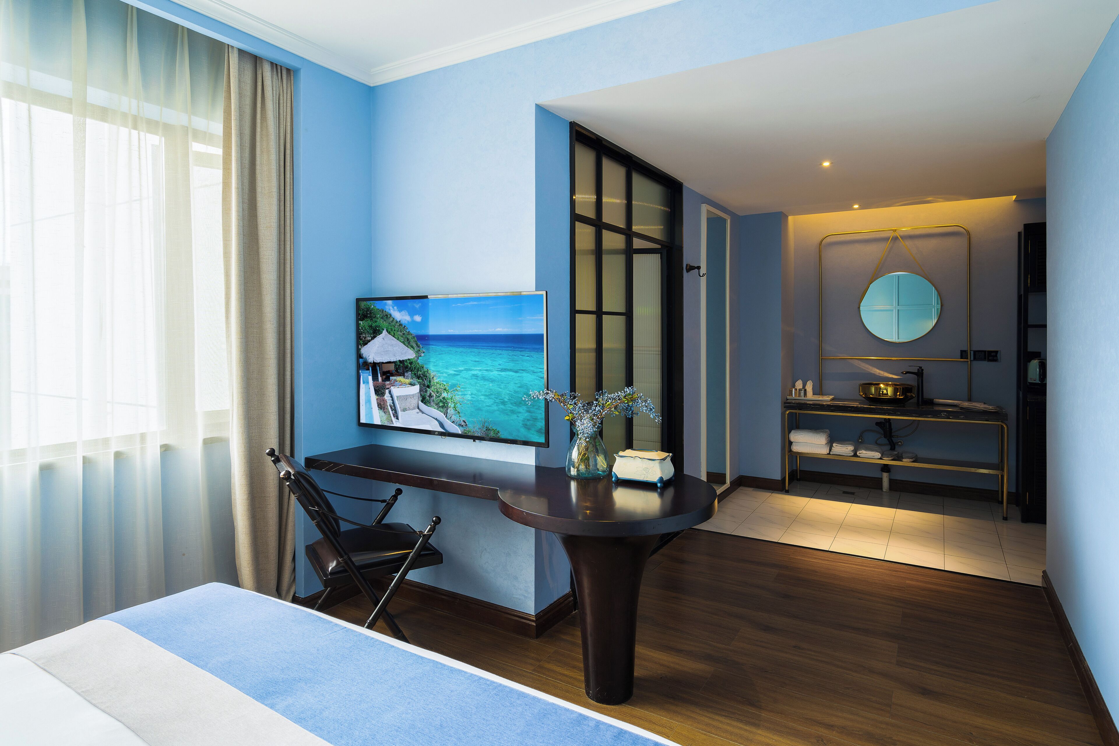 A hotel room with a bed, a desk, a chair, a TV, a window with curtains, a painting of a beach, a round mirror, and a small table with a vase.