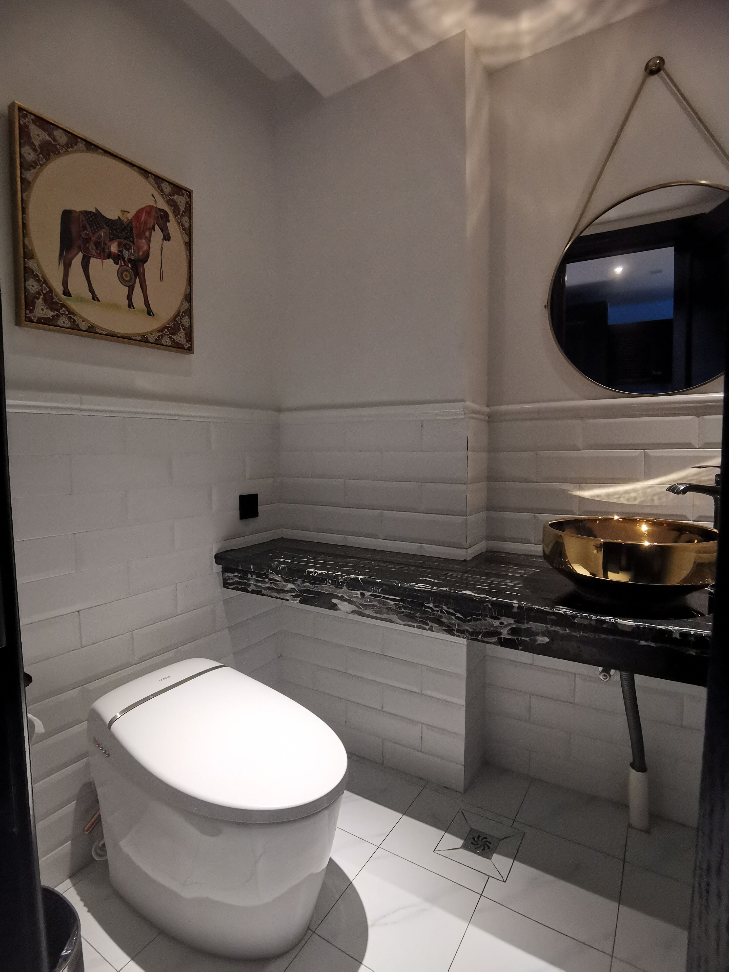 Un baño con inodoro blanco, encimera de mármol negro, un lavabo dorado, un espejo montado en la pared y un cuadro con caballos.