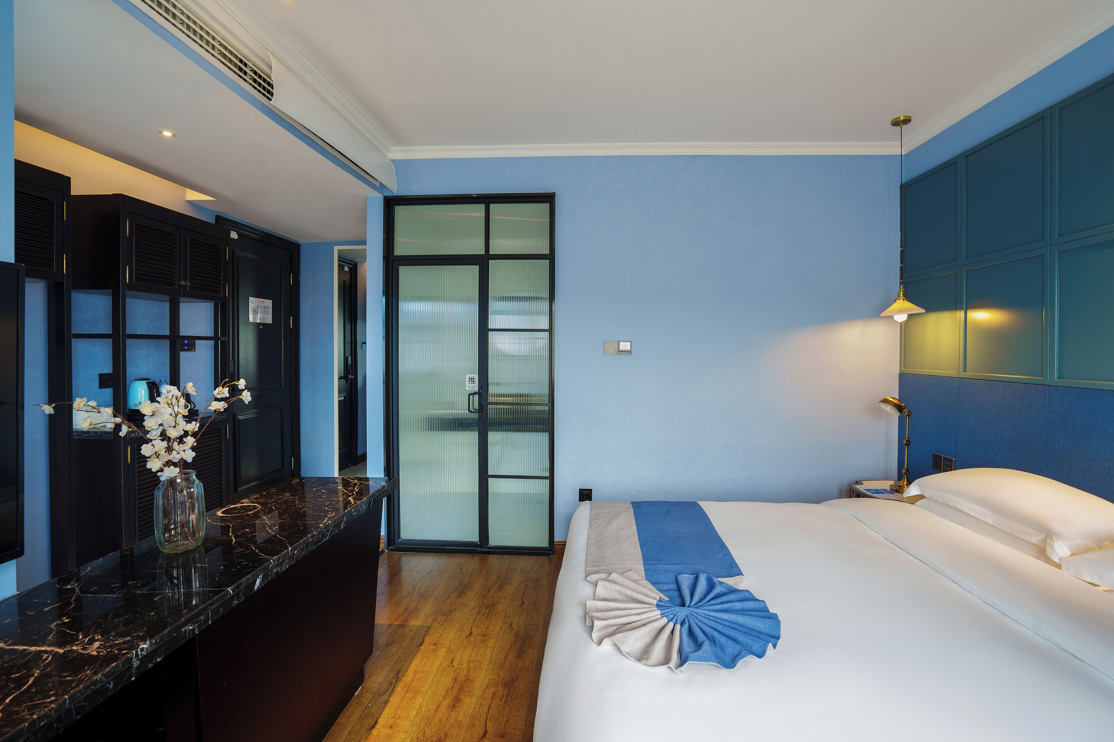 A hotel room with a bed, a desk with a vase of flowers, a nightstand with a lamp, and a sliding door to the bathroom.