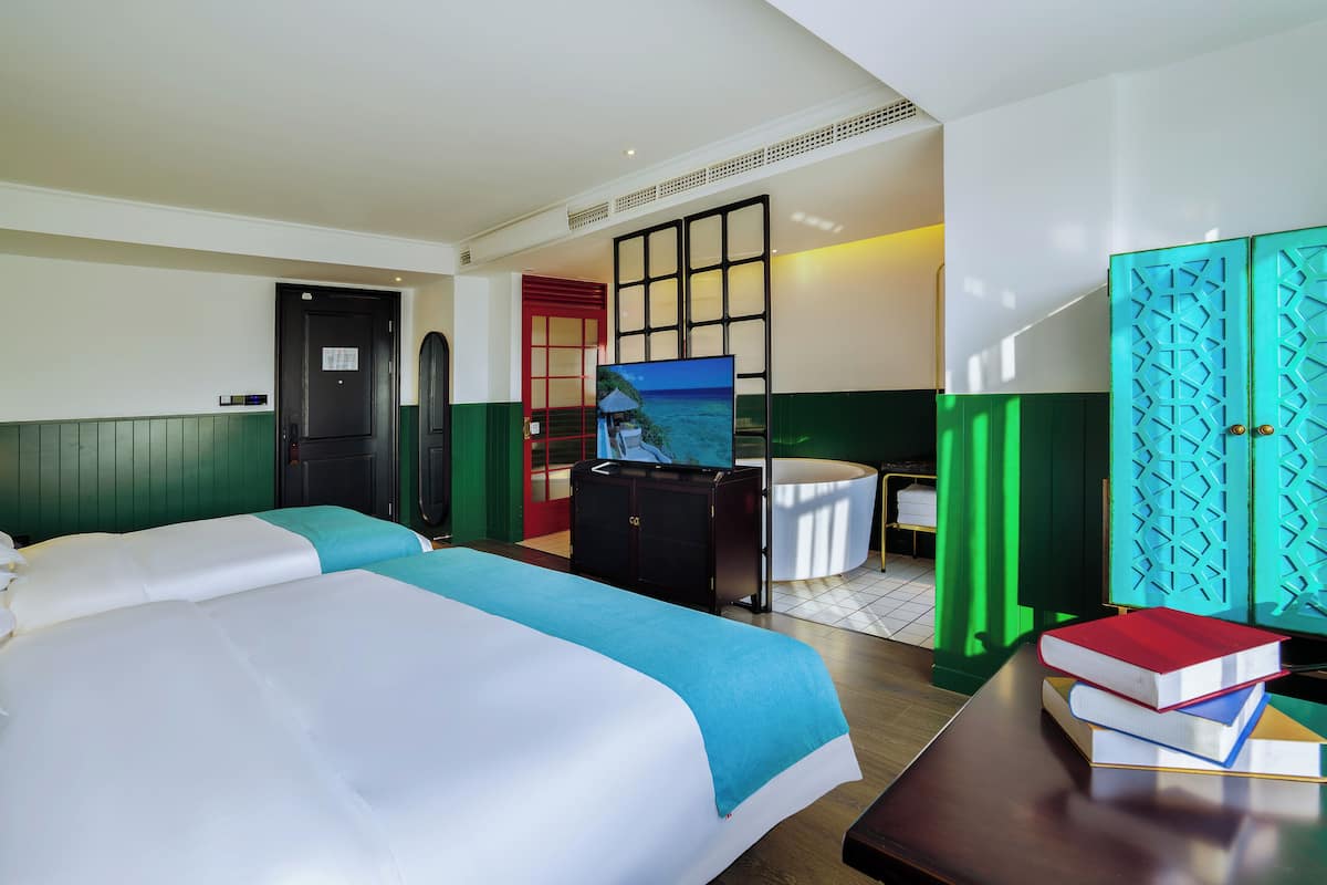 Habitación de hotel con dos camas, televisión, escritorio con libros y bañera, con una decoración en tonos verde y blanco.