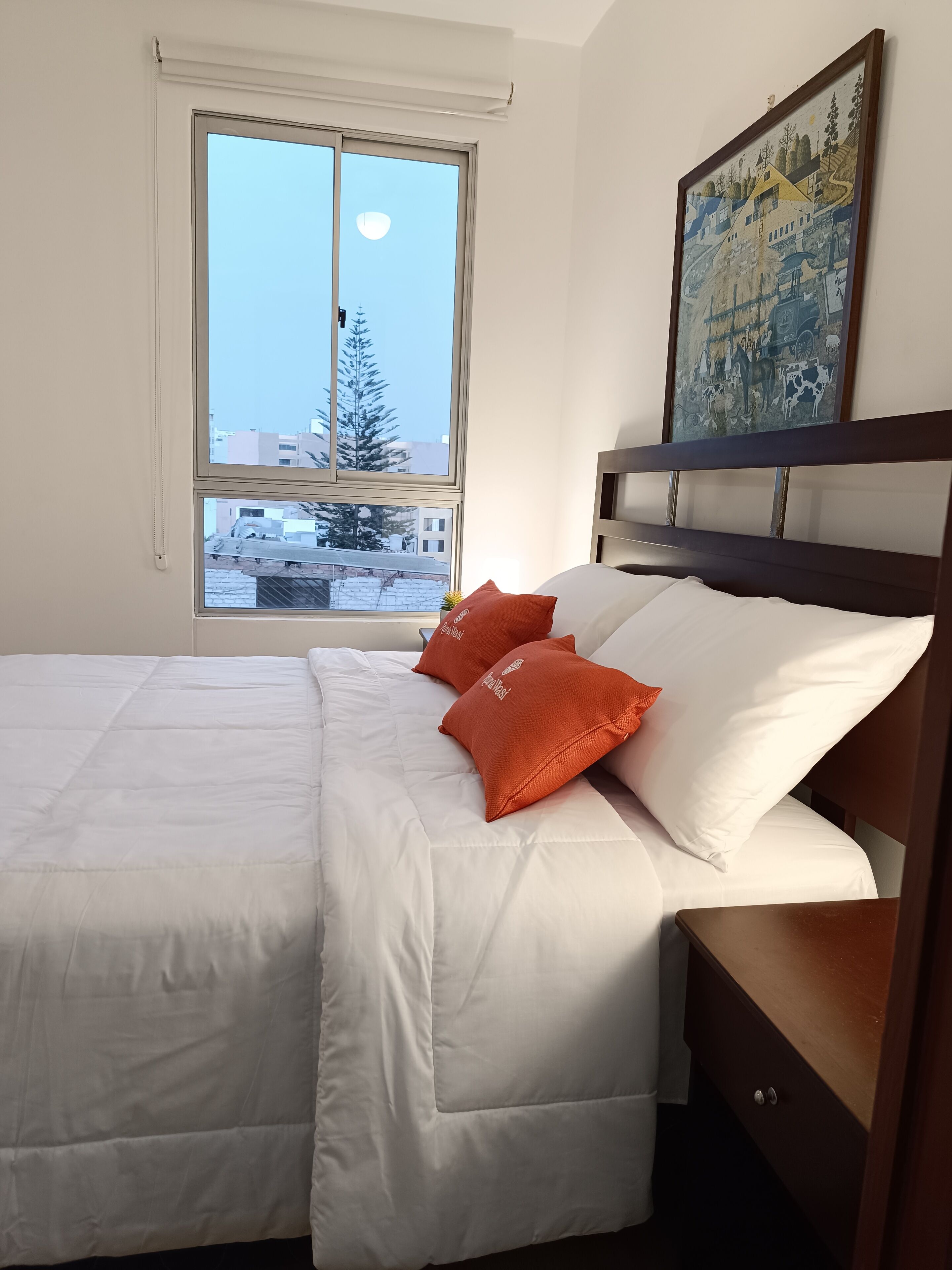 A bedroom with a large window, a bed with white bedding and orange pillows, a wooden headboard, and a painting on the wall.