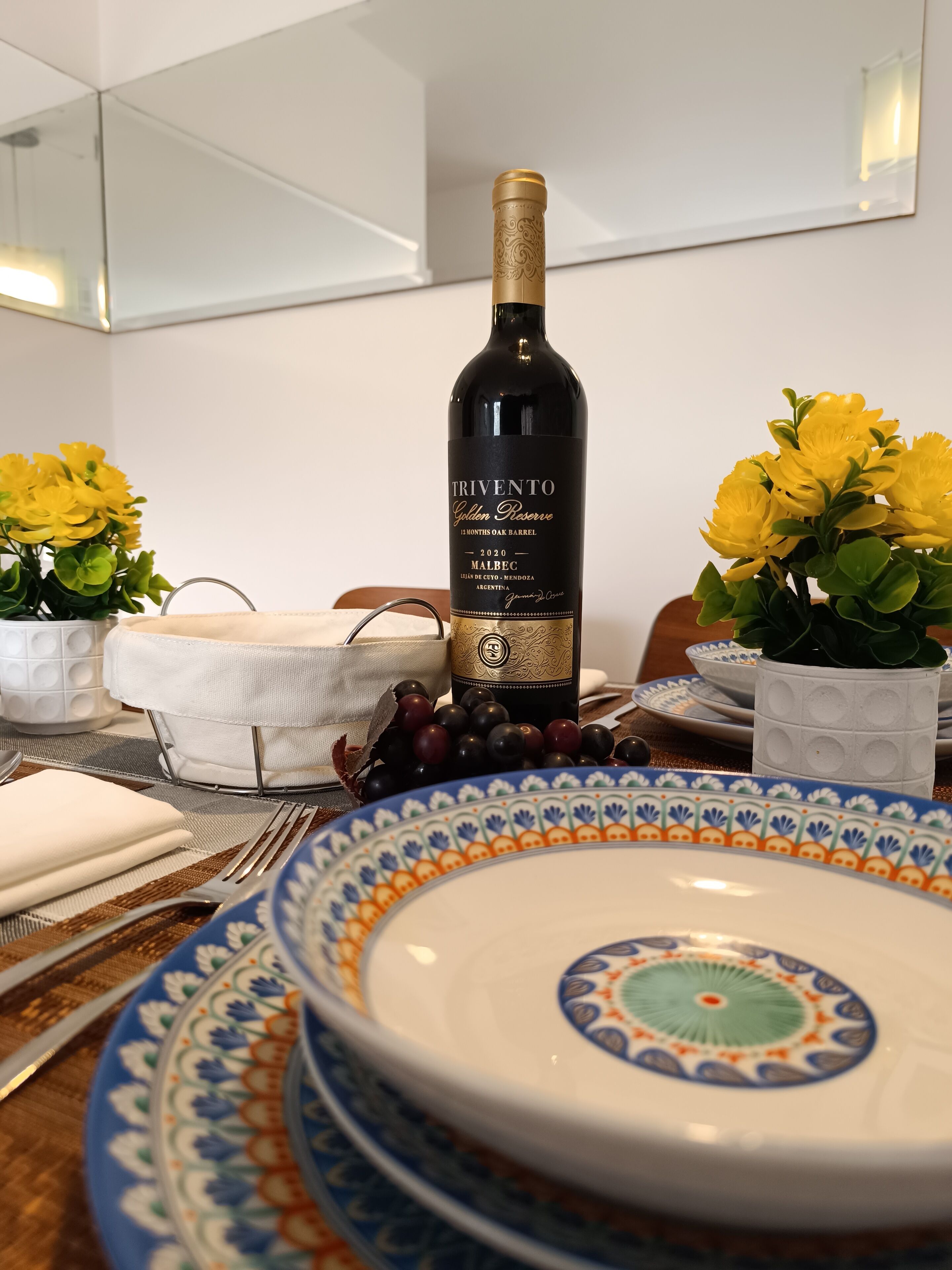 A bottle of Trivento Golden Reserve wine, a white tablecloth, a blue and orange patterned plate, and yellow flowers in a white pot.