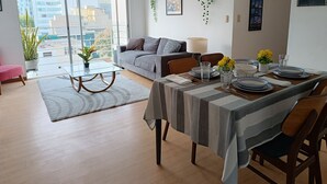 A modern living room with a dining area, large windows, a grey sofa, a coffee table, and a dining table set with plates and glasses.