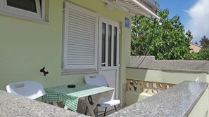 A small outdoor patio with a table and chairs, a window with shutters, and a view of a tree and building in the background. - Studio near the Old Town (Krk)