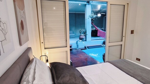 Quarto com cama, mesa de cabeceira e janela. Tem vista para uma varanda com rede e piscina.