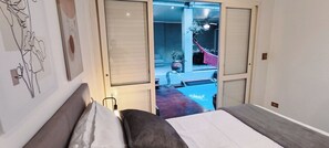 Un dormitorio con cama, mesita de noche y ventana. Se ve un balcón con hamaca y piscina.