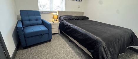 Une petite chambre avec un lit simple, un fauteuil bleu, une table de chevet avec une lampe et une fenêtre avec des stores.
