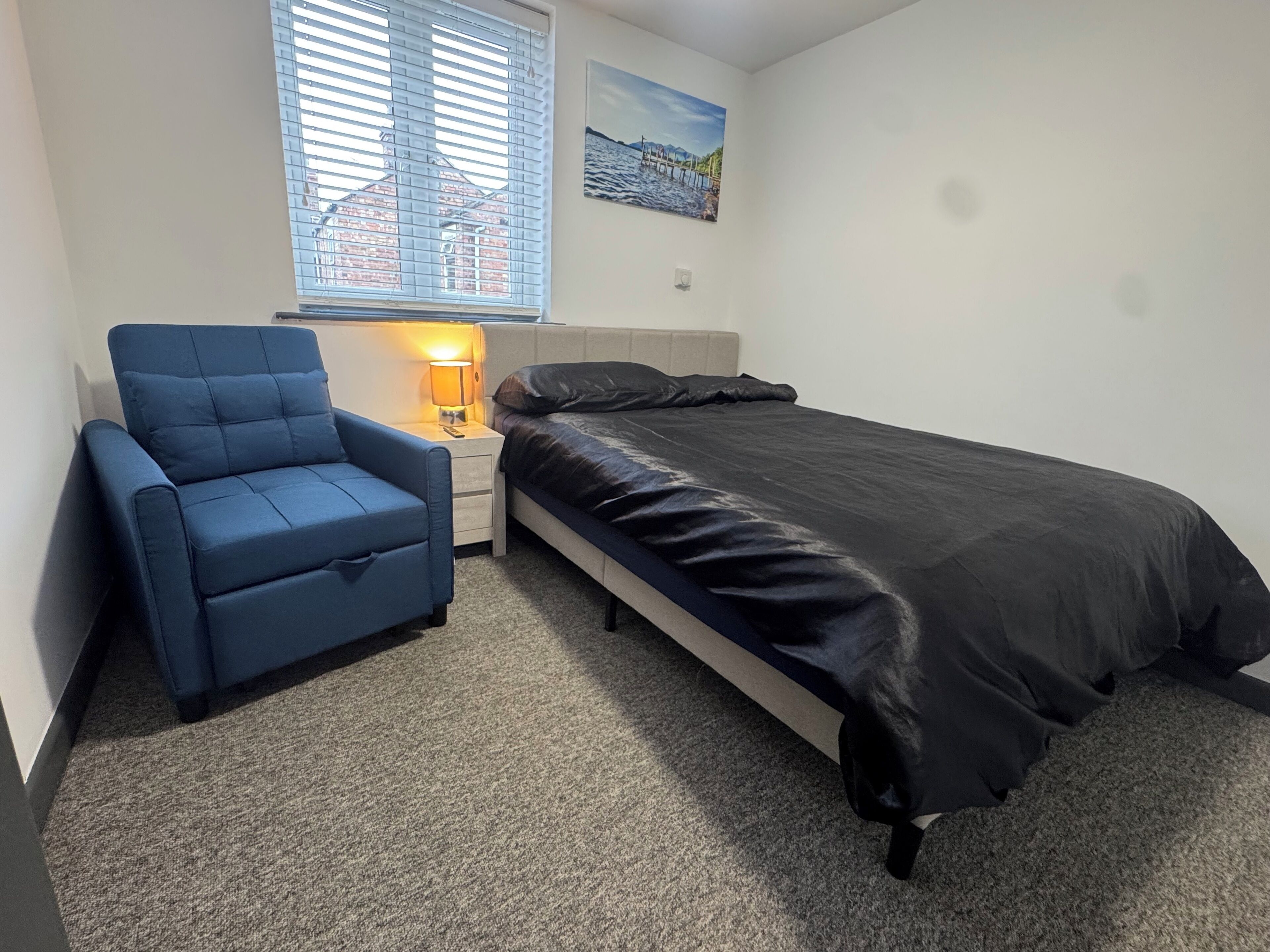 Un dormitorio pequeño y ordenado con una cama individual, una silla, una mesita de noche con lámpara y una ventana con persianas.