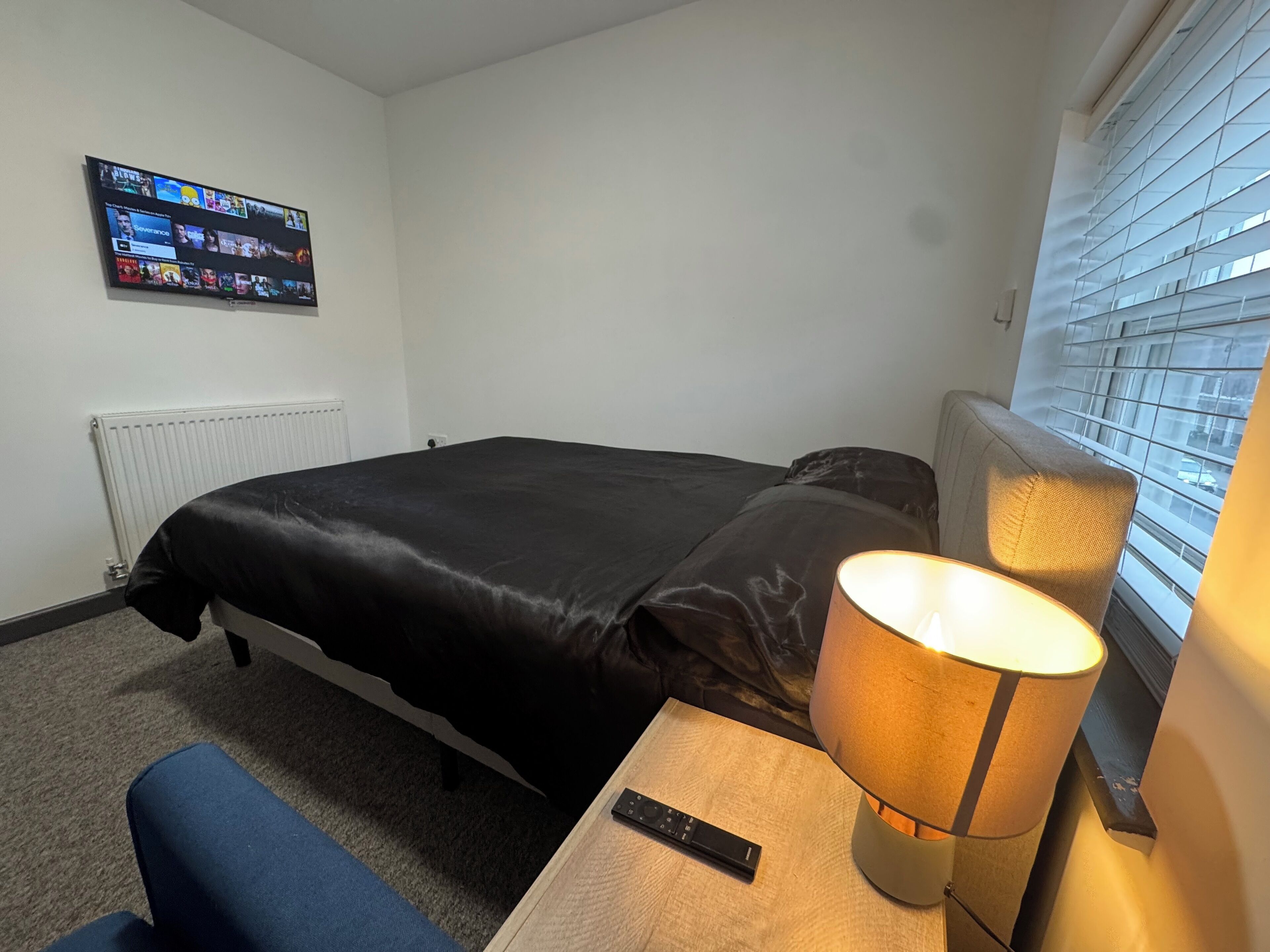 Une chambre d’hôtel avec un lit, un téléviseur fixé au mur, une lampe sur une table de chevet et une fenêtre avec des stores.