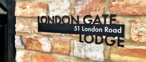 Ein schwarzes Metallschild mit der Aufschrift "LONDON GATE LODGE" und "51 London Road" an einer Ziegelwand.