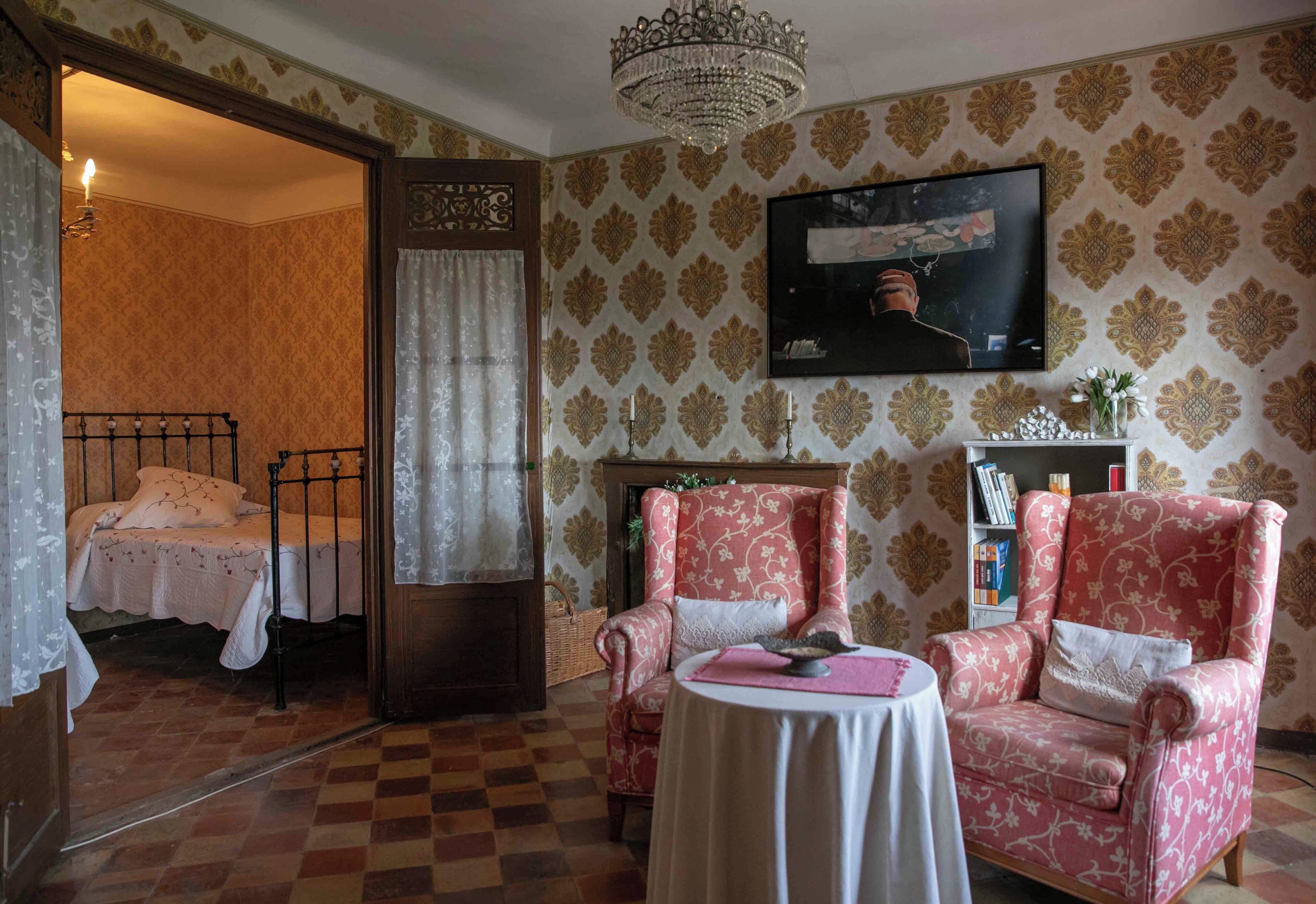 A room with a checkered floor, a bed with a white canopy, a pink armchair, a small round table, and a television mounted on the wall.