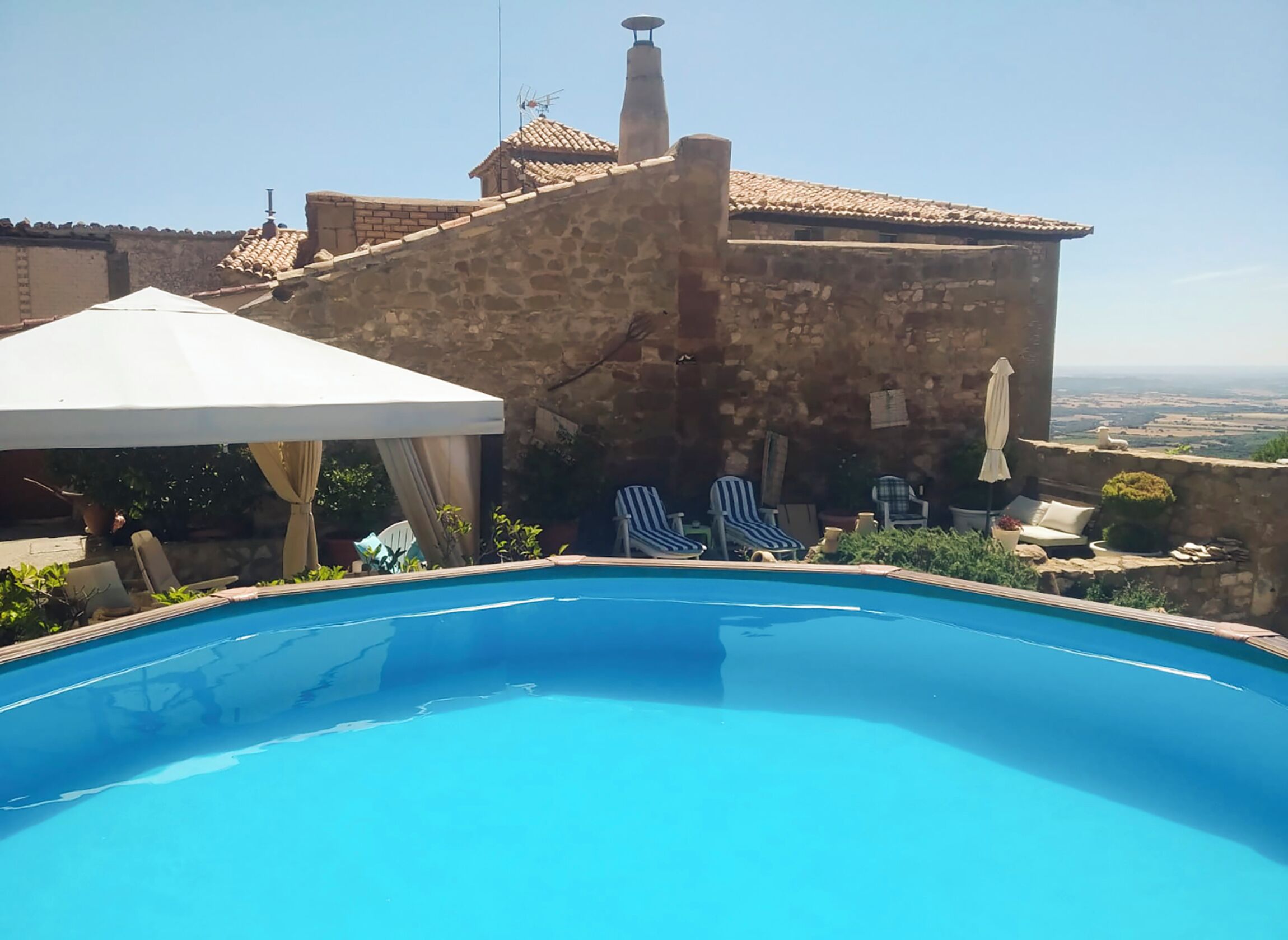A swimming pool with lounge chairs and an umbrella, a stone building with a tiled roof, and a view of the landscape.