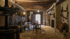 Un interior rústico con suelo de piedra, vigas de madera, una gran silla de madera y una chimenea.