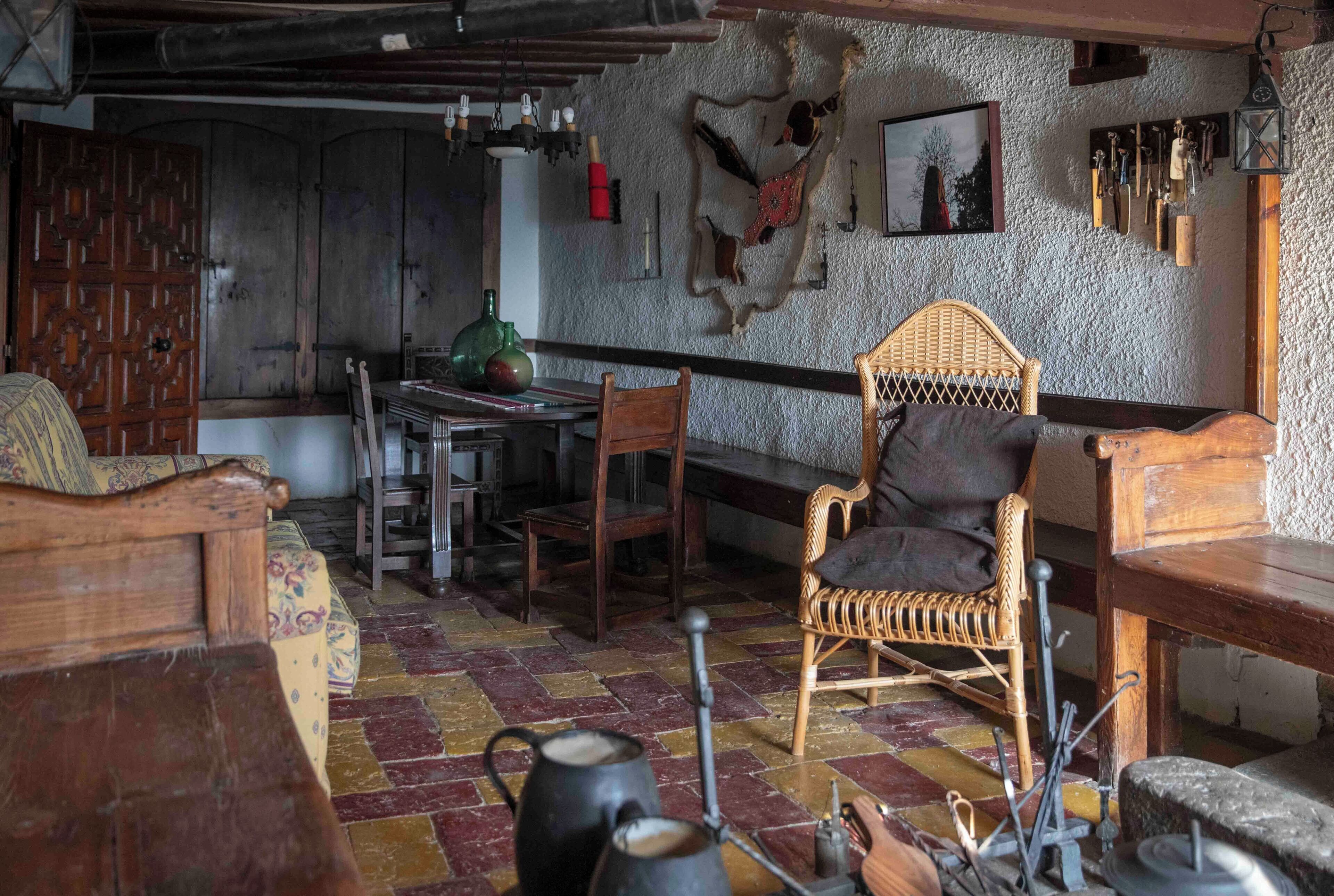 A rustic interior with a wooden dining table, chairs, a woven armchair, and a patterned sofa.
