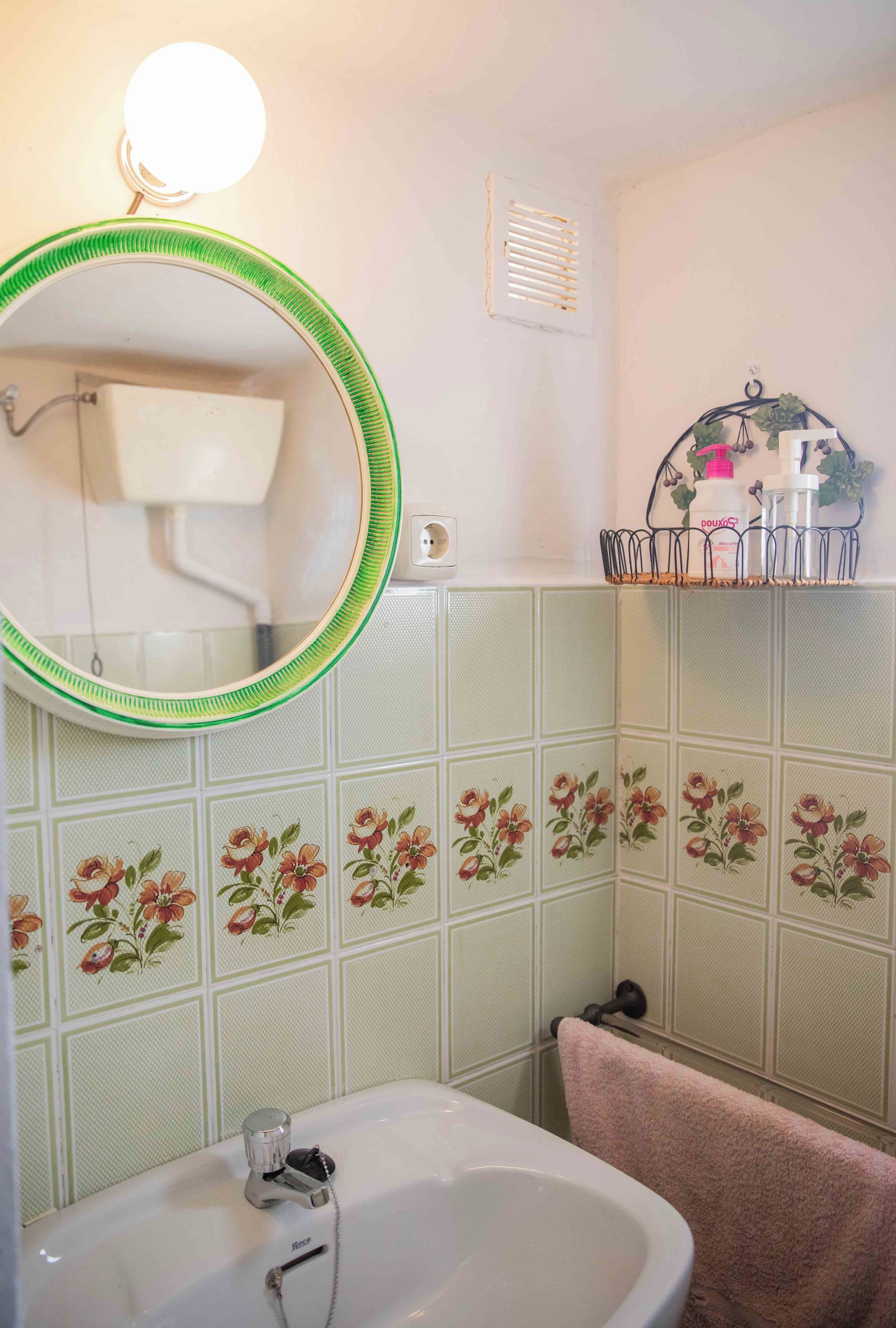 Un baño con un lavamanos blanco, espejo con marco verde, azulejos estampados con motivos florales, un perchero con artículos de aseo y una lámpara de pared.