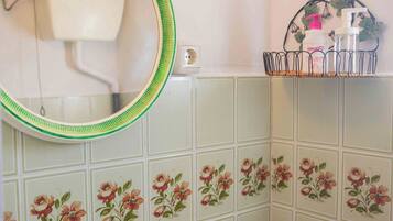 Un baño con un lavamanos blanco, espejo con marco verde, azulejos estampados con motivos florales, un perchero con artículos de aseo y una lámpara de pared.