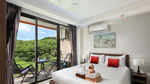 A hotel room with a large bed, two red pillows, a painting, an air conditioning unit, and a balcony with a table and chairs. - Naiharn Condominium 1 bedroom 36 sq. m. (Rawai)