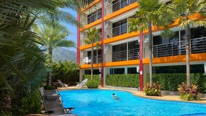 A swimming pool with lounge chairs and palm trees, surrounded by a multi-story building with orange and white exterior walls. - Naiharn Condominium 1 bedroom 36 sq. m. (Rawai)