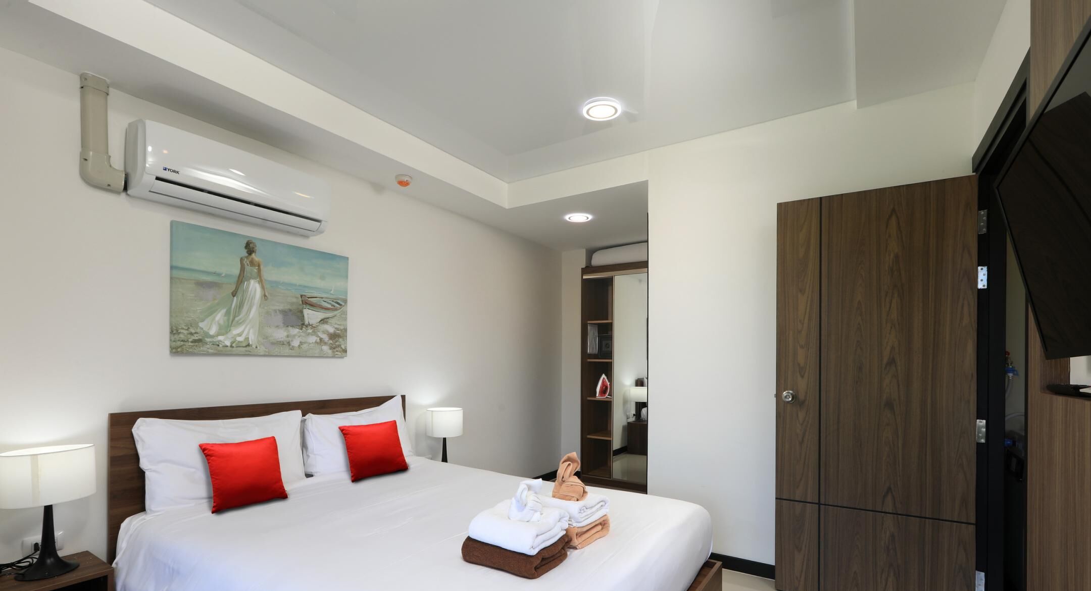 A hotel room with a bed, white bedding, red pillows, a painting, an air conditioning unit, a wardrobe, and a mirror.