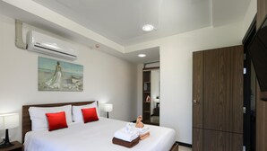 A hotel room with a bed, white bedding, red pillows, a painting, an air conditioning unit, a wardrobe, and a mirror. - Naiharn Condominium 1 bedroom 36 sq. m. (Rawai)