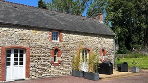 Un edificio de piedra con una puerta blanca, dos ventanas y un pequeño porche con mobiliario exterior.