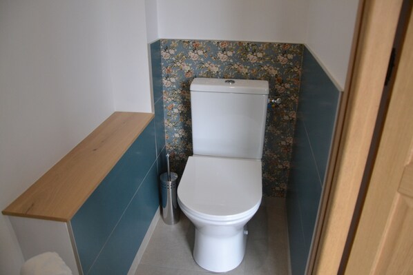 A small bathroom with a toilet, a wooden shelf, and a trash bin. - A quiet, welcoming house for 4 people, 15 minutes from Utah Beach (Picauville)