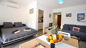Habitación de hotel moderna con cama, sofá, mesa de comedor con fruta y una kitchenette al fondo.