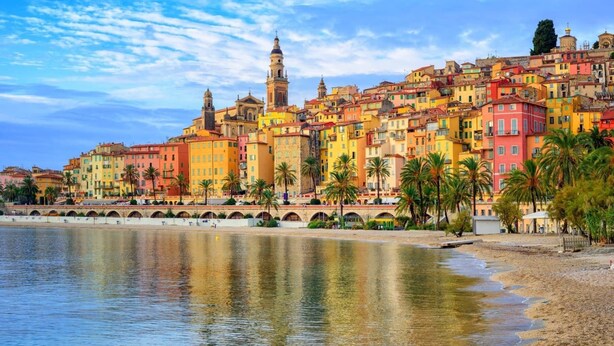 Un paisaje urbano costero con edificios coloridos, una imponente torre de iglesia y una playa con palmeras.