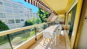 Un balcón con mesa y sillas, baranda de vidrio y vistas a edificios y follaje.