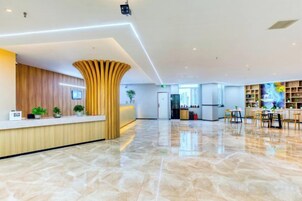 A hotel lobby with a reception desk, a wooden accent wall, a large open space, and a seating area with tables and chairs.